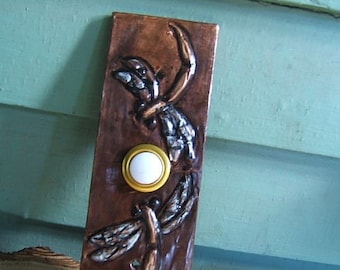 May include: A copper-colored doorbell with a white button and two dragonfly designs. The dragonfly designs are embossed and have a silver finish.
