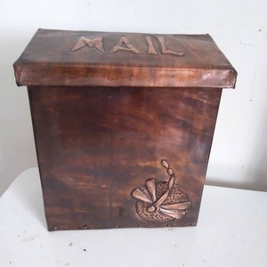 May include: A handcrafted copper mailbox with a hinged lid. The word "MAIL" is embossed on the top. A dragonfly design is featured on the front. The mailbox has a rustic, aged appearance.