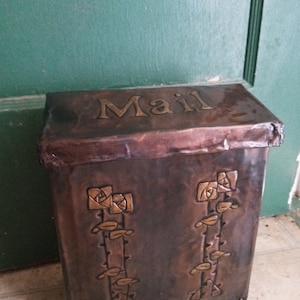 May include: A copper mailbox with a hinged lid and the word "Mail" embossed on the top. The mailbox has a decorative design of stylized roses and leaves on the front.