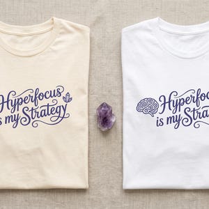 Puede incluir: Dos camisetas de manga corta, una color crema y otra blanca, cada una con un estampado gráfico de color morado oscuro de un cerebro, un cristal y el texto "Hyperfocus is my Strategy" en letra cursiva, junto a ramas de lavanda seca.