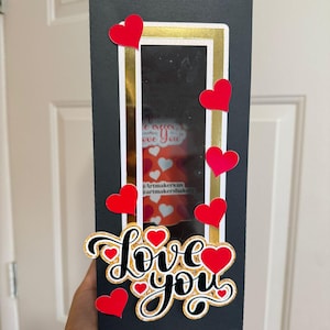 Peut inclure: Une boîte cadeau rectangulaire noire avec un bord doré et une fenêtre transparente. Des découpes en forme de cœur rouge et les mots "Love You" en cursive avec des accents de paillettes dorées sont présentés sur le devant.