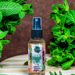 May include: A clear glass bottle with a black spray top filled with a light yellow liquid. The bottle has a label with the text "MS CHY Hydrating Rosemary Mint Mist INFUSED WITH CLOVES 4 FL OZ | 118 ML". The bottle is surrounded by green leafy plants.