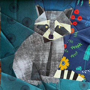 May include: A fabric art piece with a geometric raccoon design. The raccoon is mainly grey with black and white details, set against a teal and navy blue patterned background. The artwork is framed in a wooden square.