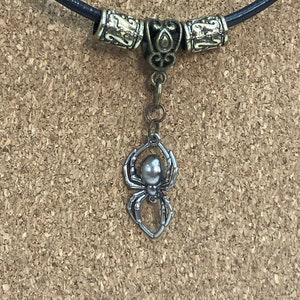 May include: A black leather necklace with a silver spider pendant. The necklace has two decorative metal beads on either side of the pendant.