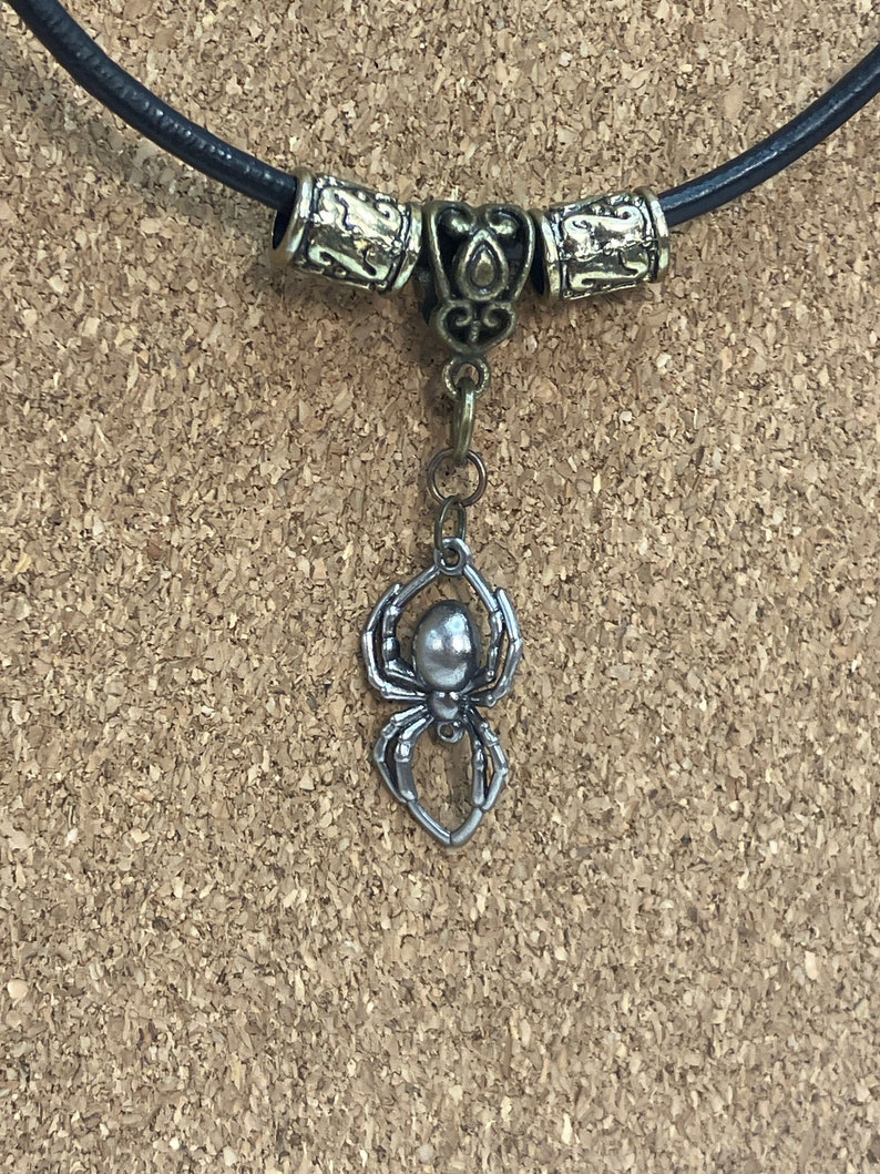 May include: A black leather necklace with a silver spider pendant. The necklace has two decorative metal beads on either side of the pendant.