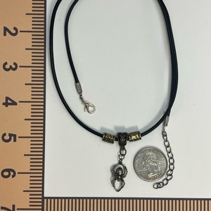 May include: A black leather cord necklace with a silver spider charm and gold-toned beads. The necklace is 18 inches long and has a lobster clasp closure.