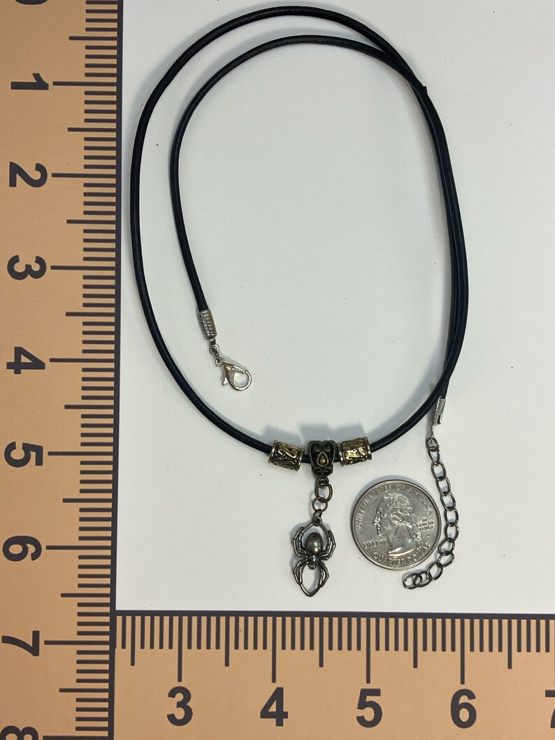 May include: A black leather cord necklace with a silver spider charm and gold-toned beads. The necklace is 18 inches long and has a lobster clasp closure.