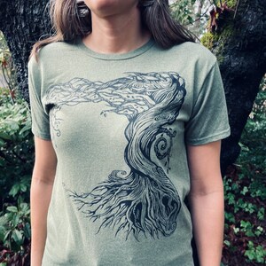 Puede incluir: Camiseta verde oliva con un gr&aacute;fico negro de un &aacute;rbol estilizado. El dise&ntilde;o del &aacute;rbol tiene ramas y ra&iacute;ces detalladas, creando una est&eacute;tica inspirada en la naturaleza. La camiseta est&aacute; hecha de un material suave.