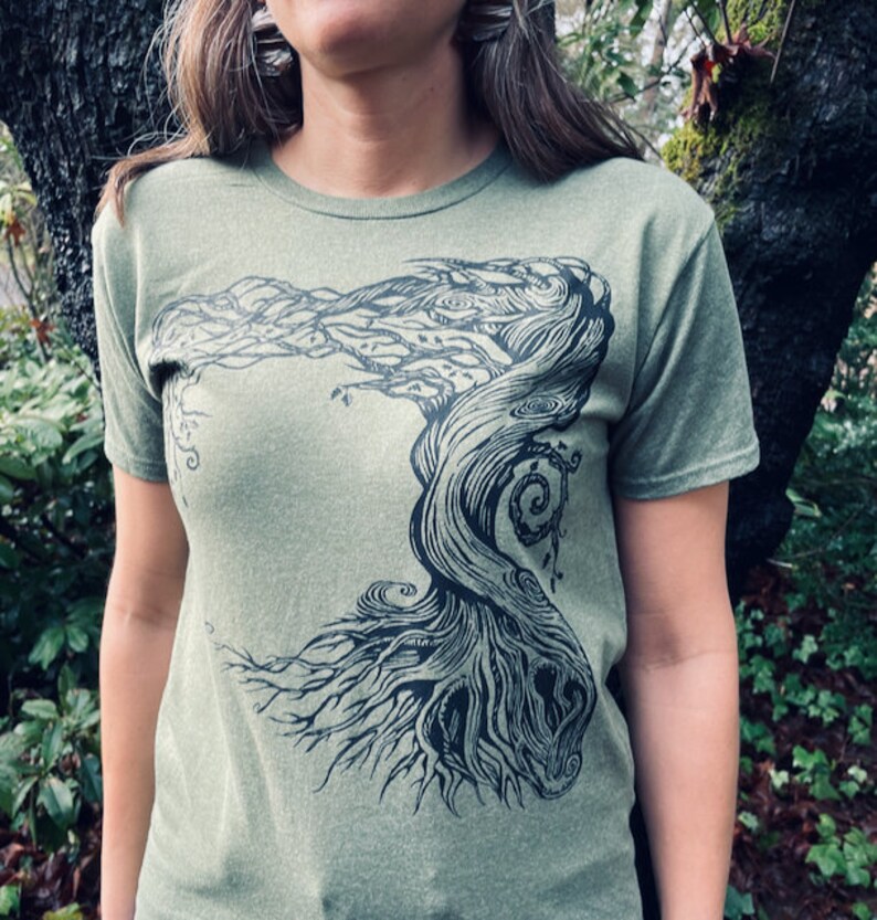 Puede incluir: Camiseta verde oliva con un gr&aacute;fico negro de un &aacute;rbol estilizado. El dise&ntilde;o del &aacute;rbol tiene ramas y ra&iacute;ces detalladas, creando una est&eacute;tica inspirada en la naturaleza. La camiseta est&aacute; hecha de un material suave.