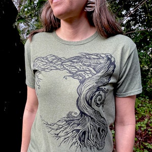 Puede incluir: Camiseta verde oliva con un gr&aacute;fico negro detallado de un &aacute;rbol estilizado. El dise&ntilde;o del &aacute;rbol tiene intrincadas ramas y ra&iacute;ces, creando una est&eacute;tica inspirada en la naturaleza. La camiseta est&aacute; hecha de una tela suave y jaspeada.