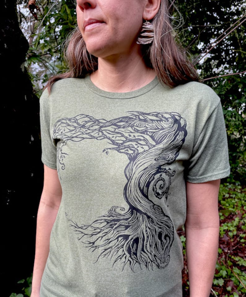Puede incluir: Camiseta verde oliva con un gr&aacute;fico negro detallado de un &aacute;rbol estilizado. El dise&ntilde;o del &aacute;rbol tiene intrincadas ramas y ra&iacute;ces, creando una est&eacute;tica inspirada en la naturaleza. La camiseta est&aacute; hecha de una tela suave y jaspeada.