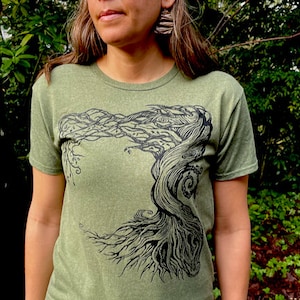 Puede incluir: Camiseta verde oliva con un dise&ntilde;o de &aacute;rbol negro. El dise&ntilde;o del &aacute;rbol presenta un tronco y ramas detallados y arremolinados. La camiseta de manga corta y cuello redondo est&aacute; hecha de un material suave.