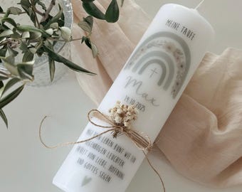 May include: White pillar candle with a rainbow design, the text "MEINE TAUFE", and the name "Max" with a date. The candle is decorated with a small bundle of dried flowers and tied with twine. The text "AUF DEN LEBEN BUNT WIE EIN REGENBOGEN LEUCHTEN..." is printed on the candle.