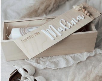 May include: A light wooden box with a lid bearing the name "Malina" in white script. Inside, a candle with a rainbow design and a pair of white baby shoes. The lid is decorated with small wooden beads and dried flowers.