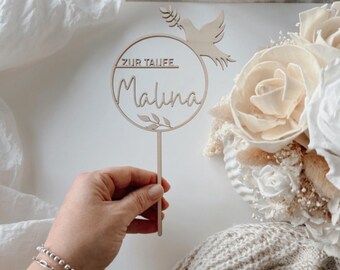 May include: A wooden cake topper with the name "Malina" in script, encircled by a ring with the text "ZUR TAUFE" above. A dove and leaves are cut out of the wood. The topper is held by a person.
