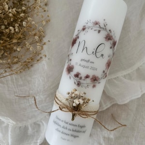 Może przedstawiać: Biały świeca kolumnowa ozdobiona kwiatowym wieńcem i imieniem "Mila". Świeca zawiera datę "11. August 2024" i werset biblijny. U podstawy znajduje się wstążka z juty i suszone kwiaty.