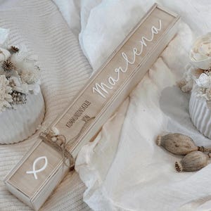 Peut inclure: Une boîte en bois de couleur claire avec le nom "Marlena" en écriture blanche. La boîte est décorée d'un symbole de poisson blanc et des mots "Kommunionskerze von". La boîte est attachée avec de la ficelle et décorée de fleurs séchées.
