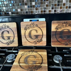 May include: Four wooden chopping boards with a floral design and the letter "G" engraved in the centre. The boards are arranged in a row on a black hob.