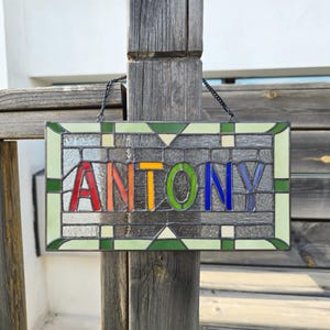 May include: A stained glass window panel featuring the name "ANTONY" in colourful letters. The letters are outlined in black and the background is a mosaic of small, clear glass pieces.