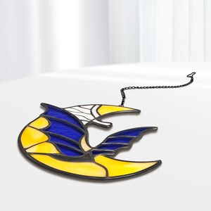 May include: A stained glass suncatcher in the shape of a stylized bird or bat. The design features yellow, blue, and white glass pieces, outlined with black metal. A black chain is attached for hanging.