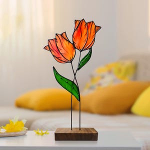 Pode incluir: Uma escultura de vidro colorido que representa duas tulipas laranja com folhas verdes sobre uma base de madeira.