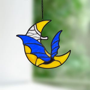 May include: A stained glass bird suncatcher with a yellow and blue design. The bird is hanging from a black chain. The bird's wings and body are yellow, with blue and white accents. The suncatcher is a decorative item.
