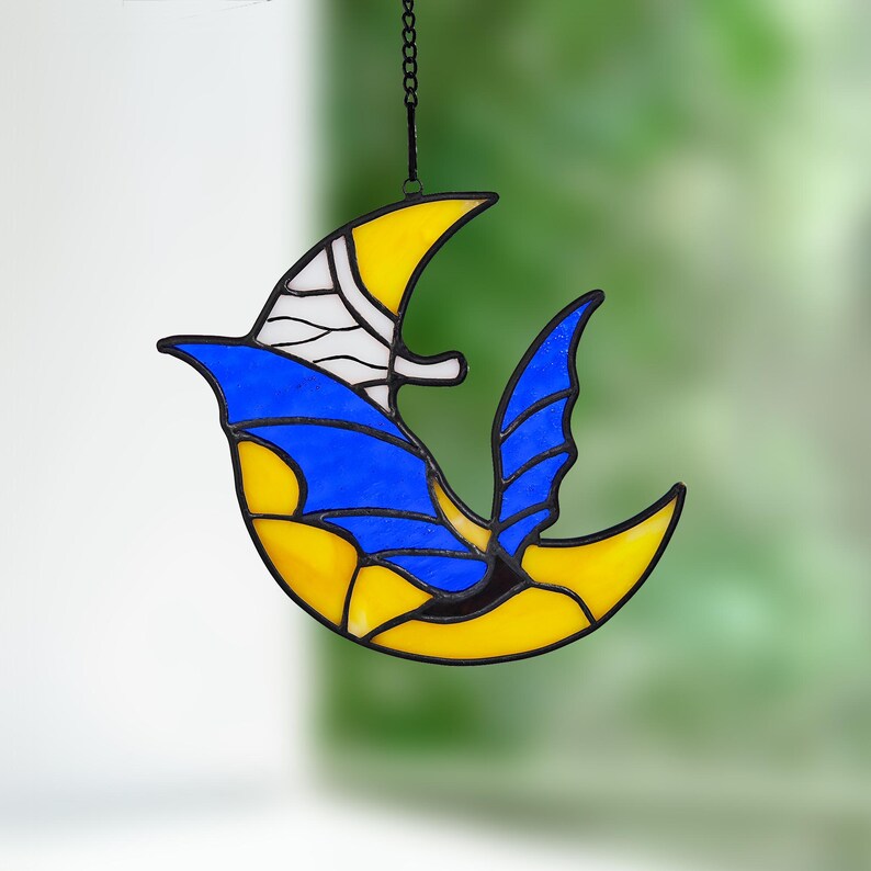 May include: A stained glass bird suncatcher with a yellow and blue design. The bird is hanging from a black chain. The bird's wings and body are yellow, with blue and white accents. The suncatcher is a decorative item.