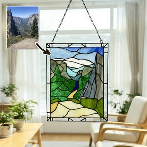 Könnte beinhalten: Buntglasfenster mit einer Berglandschaft in Blau-, Grün- und Grautönen. Das rechteckige Panel hat einen schwarzen Metallrahmen und wird von einer Kette gehalten. Ein kleineres Bild der Landschaft befindet sich in der oberen linken Ecke.