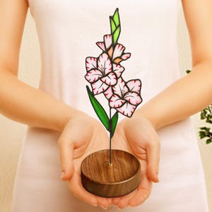 以下が含まれることがあります： 木製のスタンドに置かれたピンクと白のステンドグラスのグラジオラスの花。花はガラス製で、繊細で複雑なデザインが施されています。