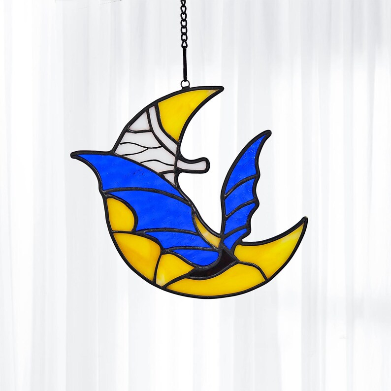 May include: Stained glass bird suncatcher in yellow, blue, and white. The bird is suspended by a black chain. The bird's wings and body are yellow and blue, with white accents. The suncatcher is designed to hang in a window.