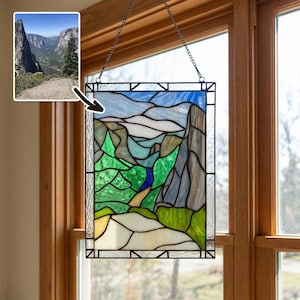 May include: A rectangular stained glass panel depicting a mountain landscape with a blue sky, grey rock formations, green trees, and a light-colored path. The piece is framed in black metal and hangs from a thin metal chain.