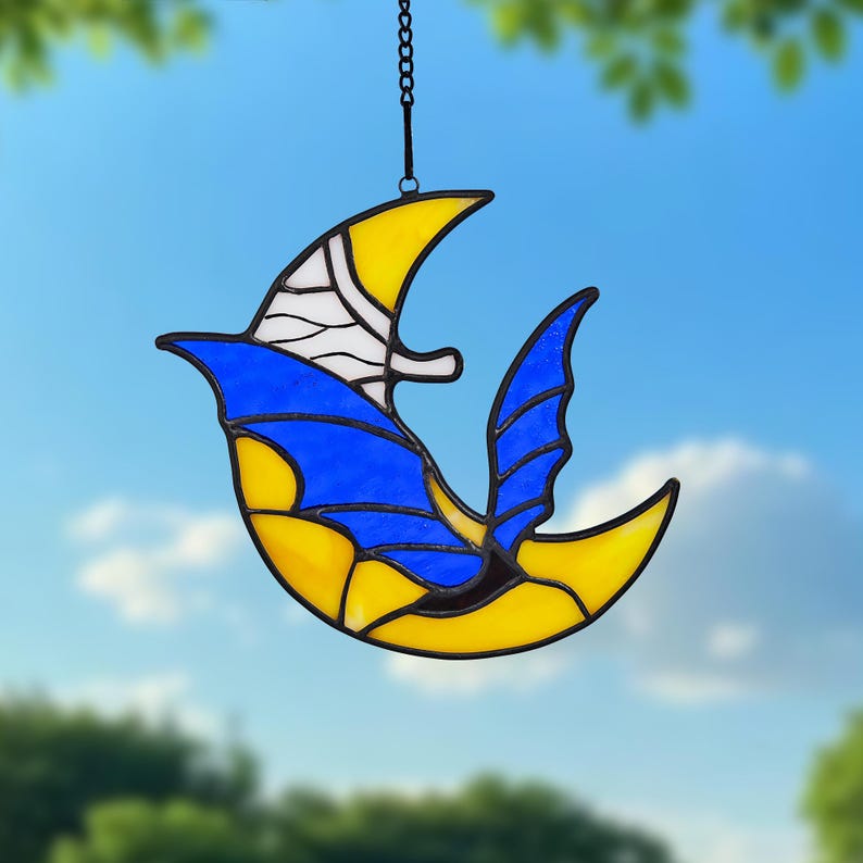May include: A stained glass suncatcher in the shape of a bird or crescent moon. The design features yellow, blue, and white glass pieces, with a black metal outline and chain for hanging. The suncatcher is suspended against a blue sky.