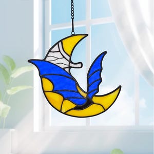 May include: Stained glass suncatcher in the shape of a bird, featuring yellow, blue, and white sections. The bird is suspended from a black chain, designed for window or wall display. The design is reminiscent of a bird in flight.