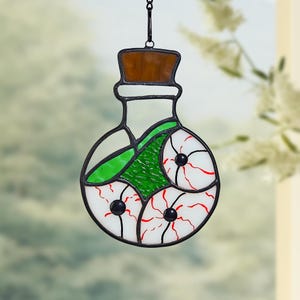 May include: Stained glass potion bottle with a brown cork and black outline. The bottle contains three white eyeballs with red veins and black pupils, and green textured glass. A chain is attached for hanging.