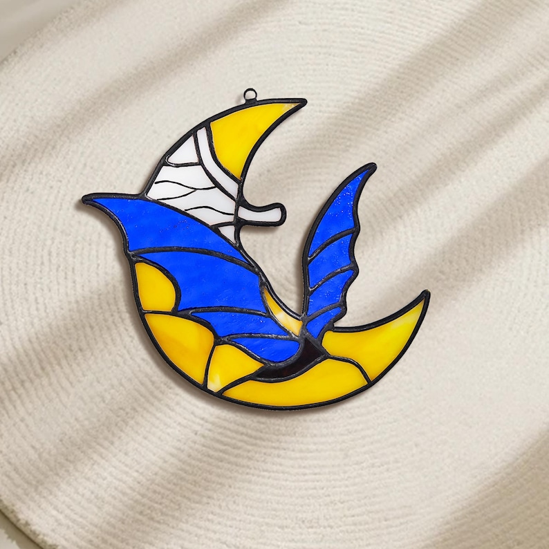 May include: A stained glass bird-shaped suncatcher with a black outline. The bird features yellow, blue, and white glass pieces. The suncatcher has a loop for hanging and is set against a neutral background.