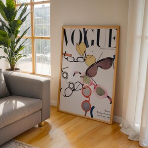 May include: A framed vertical poster featuring a vintage Vogue magazine cover from August 1936. The artwork displays a white background with the black text VOGUE at the top and various colorful illustrations of eyeglasses and sunglasses scattered throughout.