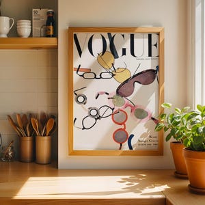 May include: A framed Vogue magazine cover print from August 1996 featuring a white background with various colorful illustrations of eyeglasses and sunglasses scattered across the page. The frame is made of light-toned wood and hangs on a wall.