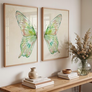Puede incluir: Dos cuadros de pared enmarcados con un diseño de ala de mariposa dividida en tonos acuarela de verde claro, rosa suave y crema. Cada impresión rectangular está encerrada en un marco de madera clara, mostrando un ala de mariposa simétrica.