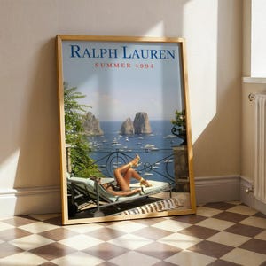 May include: A framed vertical poster titled RALPH LAUREN SUMMER 1994 featuring a coastal scene with a person reclining on a lounge chair on a balcony overlooking the sea, rocky cliffs, and boats. The poster is encased in a light wood frame.