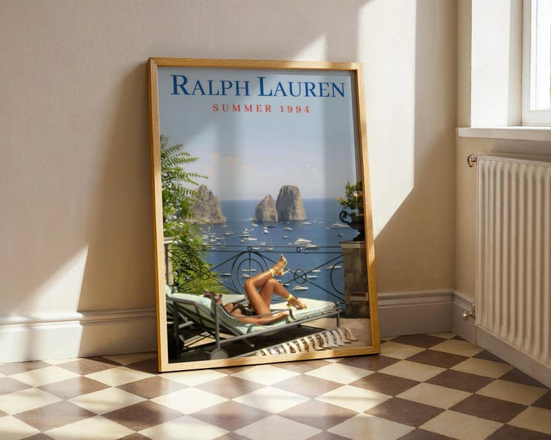 May include: A framed vertical poster titled RALPH LAUREN SUMMER 1994 featuring a coastal scene with a person reclining on a lounge chair on a balcony overlooking the sea, rocky cliffs, and boats. The poster is encased in a light wood frame.