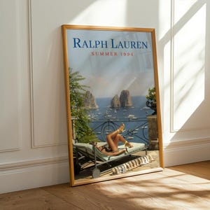 May include: A framed poster featuring the text RALPH LAUREN SUMMER 1994. The image depicts a person reclining on a lounge chair on a balcony overlooking a blue sea with rocky islands and boats. A row of sandals is lined up on the floor nearby.