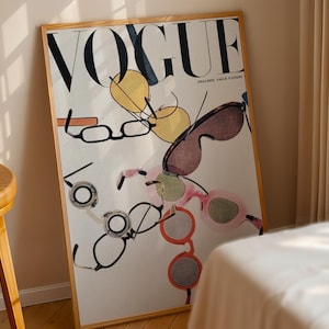 May include: A framed wall art print featuring the word VOGUE in large black letters at the top. The design displays a scattered collection of various eyeglasses and sunglasses in shades of yellow, brown, pink, and orange against a white background.
