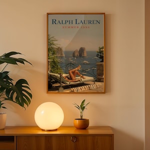 May include: A rectangular framed wall art print titled Ralph Lauren Summer 1994, featuring a coastal scene with a person lounging on a deck chair overlooking the sea, rocky cliffs, and boats, displayed in a thin wooden frame on a plain wall.