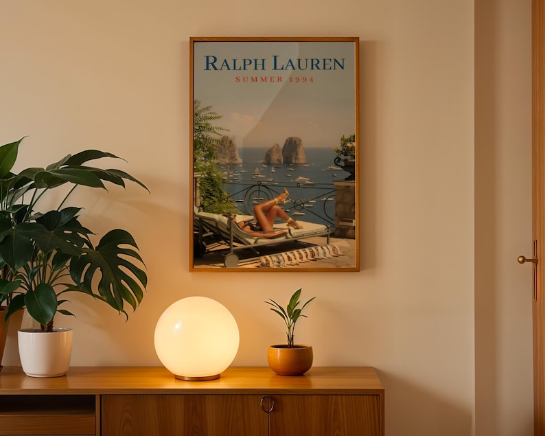May include: A rectangular framed wall art print titled Ralph Lauren Summer 1994, featuring a coastal scene with a person lounging on a deck chair overlooking the sea, rocky cliffs, and boats, displayed in a thin wooden frame on a plain wall.