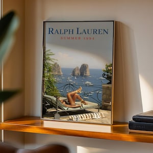 May include: A framed rectangular poster featuring the text RALPH LAUREN SUMMER 1994 at the top. The image depicts a person reclining on a lounge chair on a terrace overlooking a blue sea with rock formations, boats, and a metal railing in the distance.