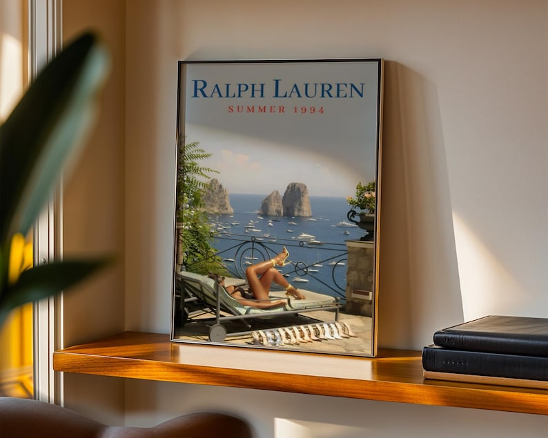 May include: A framed rectangular poster featuring the text RALPH LAUREN SUMMER 1994 at the top. The image depicts a person reclining on a lounge chair on a terrace overlooking a blue sea with rock formations, boats, and a metal railing in the distance.