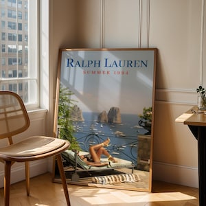 May include: A rectangular framed poster titled Ralph Lauren Summer 1994, featuring a coastal scene with a person lounging on a deck chair overlooking a sea with rock formations, boats, and a balcony railing, all rendered in a vintage photographic style.