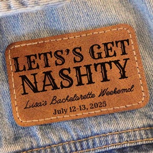 Op de afbeelding: Een bruin leren patch met de tekst "Let's Get Nashy" in zwarte letters. Onder de tekst staat "Lisa's Bachelorette Weekend" en "July 12-13, 2025" in kleinere zwarte letters.