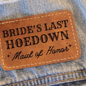 Puede incluir: Un parche de cuero marrón con el texto "Bride's Last Hoedown" y "Maid of Honor" en letras negras. El parche tiene un borde cosido y dos pequeñas estrellas.