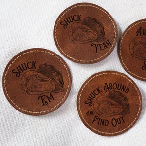 Puede incluir: Cuatro posavasos de cuero marrón con borde cosido y diseños de ostras grabados. Los posavasos presentan las frases "Shuck Em", "Shuck Around And Find Out", "Aw Shucks" y "Shuck Yeah".
